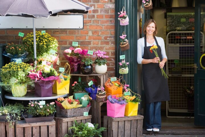 A woman floral designer standing at the door of the flower shop, holding a couple of flowers on her hand and showing at her right, bouquets of many color of flowers.